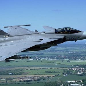 May include: A gray fighter jet soaring through a clear blue sky. The aircraft, with the number 9243 on its side, is in flight over a landscape of green fields and a distant town. The jet has a sleek, modern design.
