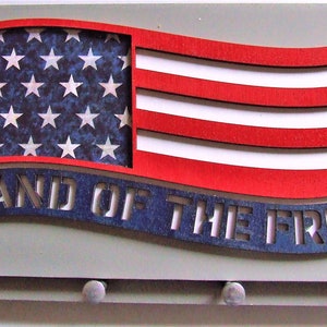 May include: A wooden key holder shaped like an American flag with the words "Land of the Free" cut out. The flag is red, white, and blue with a blue background and white stars. The key holder has five hooks on the bottom.