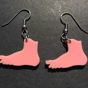 May include: A pair of dangle earrings featuring flat, light pink, foot-shaped charms made of a smooth material. Each charm is suspended from a silver-toned metal fishhook ear wire by a small jump ring, creating a simple and distinct design.