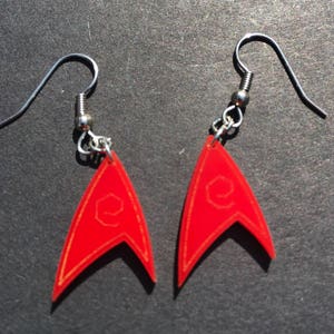 May include: A pair of red dangle earrings featuring a pointed, triangular shape with a thin gold-colored outline and a small, circular emblem in the center. The earrings are attached to silver-toned metal fishhook wires for pierced ears.