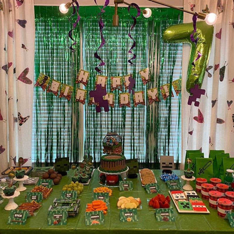 May include: A Minecraft-themed birthday party table setting with green and brown decorations. The table is decorated with a green tablecloth, green and brown paper plates, and a green and brown banner that says "Happy 7th Birthday". There are also several food items on the table, including a chocolate cake, fruit, and snacks.
