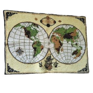 May include: A rectangular tufted rug featuring a vintage-style world map with two circular hemispheres. The continents are depicted in shades of green and brown against a cream background, with a black border and decorative corner accents.