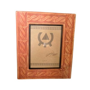 May include: A rectangular picture frame with a terracotta-colored border featuring a raised, repeating braided rope pattern. The inner display area contains a beige insert with a dark gray geometric symbol, a signature, and the text 5 X 7.