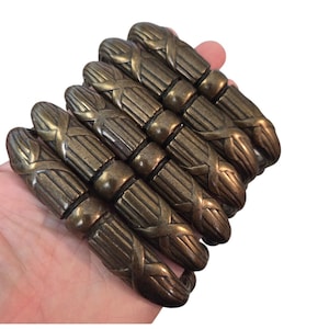 May include: A wide bracelet composed of five parallel, cylindrical metal links with a dark bronze finish. Each link features a vertical ribbed texture and a central horizontal band with a cross-shaped design, connected by small dark circular spacers.