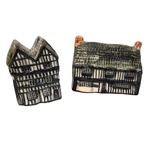 May include: Two ceramic miniature houses with black and white Tudor-style facades and dark gray roofs. The houses have small windows and are set against a white background. The roofs have a textured appearance, and the houses are detailed with architectural elements.