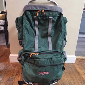 May include: A dark green JanSport external frame backpack featuring a large main compartment, a smaller front zippered pocket, and grey vertical straps. The bag includes orange zipper pulls and a silver metal frame structure at the top and bottom.