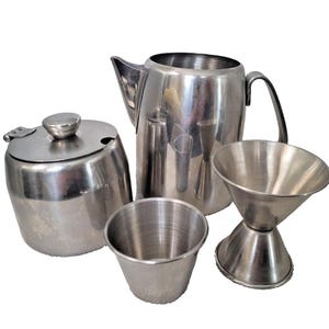 May include: A stainless steel set consisting of a lidded sugar bowl, a tall pitcher with a handle and spout, a small cylindrical cup, and a double-sided jigger. Each piece features a polished metallic finish and a smooth, simple construction.