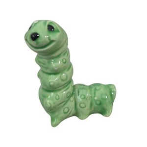 May include: A ceramic figurine of a smiling green caterpillar with black eyes and white accents. The caterpillar has a segmented body with raised dots and is posed in a curved position.