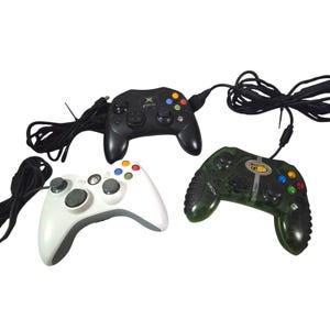 May include: Three wired game controllers for Xbox consoles. One is a black controller with a central logo, one is a white controller with gray joysticks, and one is a translucent green controller with a Mad Catz logo and black textured side grips.
