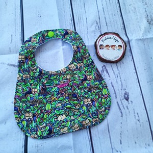 May include: A green, black and yellow baby bib with a Teenage Mutant Ninja Turtles print. The bib has a green button and a round opening for the neck. The bib is handmade in Wales by Kiddie Clips.