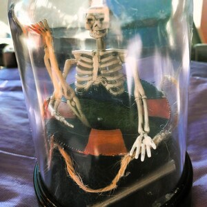 May include: A miniature skeleton figure inside a clear glass dome. The skeleton is seated on a black and orange life preserver. The dome sits on a black base. The skeleton is holding a rope.