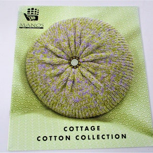 May include: A green and purple knitted cushion with a decorative button in the center. The cushion is on a green background with a wave pattern. The text "Cottage Cotton Collection" is printed below the cushion.