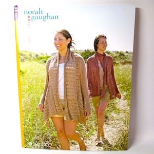 May include: Two women wearing knitted cardigans and scarves walk along a path in a grassy field. The text "norah gaughan collection" is printed on the cover of the book.