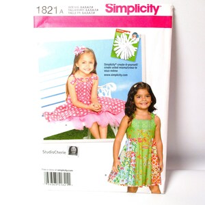 May include: Simplicity sewing pattern 1821A featuring two girls' dresses. One dress is pink with white polka dots, the other is green with floral and patterned panels. The pattern includes sizes 3, 4, 5, 6, 7, and 8.
