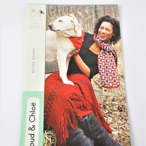 May include: A magazine cover featuring a woman and a dog. The woman is wearing a red and blue patterned scarf and holding a dog wearing a red bandana. The woman is also wearing a red blanket with fringe. The magazine cover has the text "Aztec Scarf" and "Spud & Chloe".