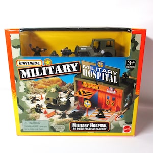 May include: A Matchbox Military Hospital 14-piece fold-up playset in a rectangular box. The packaging features a camouflage border, a clear window displaying a toy jeep and figures, and graphics of a military hospital building and soldiers.