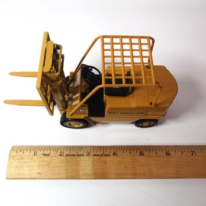 Może przedstawiać: Ż&oacute;łta zabawka w&oacute;zka widłowego Caterpillar z klatką ochronną i czarną kierownicą. W&oacute;zek widłowy ma dwa przednie widły i czarne opony. Obok w&oacute;zka widłowego umieszczono drewnianą linijkę.