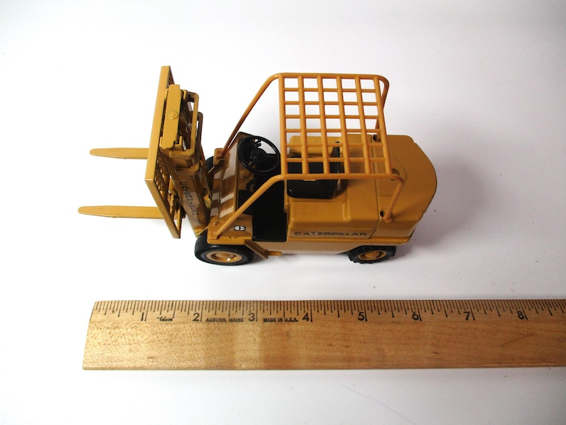Może przedstawiać: Ż&oacute;łta zabawka w&oacute;zka widłowego Caterpillar z klatką ochronną i czarną kierownicą. W&oacute;zek widłowy ma dwa przednie widły i czarne opony. Obok w&oacute;zka widłowego umieszczono drewnianą linijkę.