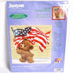 May include: A cross-stitch kit featuring a brown teddy bear holding an American flag. The kit includes 12-mesh cotton interlock canvas, wool yarn, a needle, a graph, and instructions. The finished size is 16 inches by 16 inches.