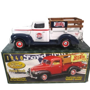 May include: A die-cast metal replica of a 1940 Ford pickup truck. The truck is white with blue fenders and a Pepsi-Cola logo. The truck sits on a box with a picture of a red truck and the words "1940 Ford Pickup Truck."