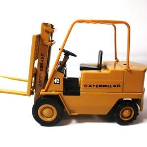 Może przedstawiać: Ż&oacute;łta zabawka w&oacute;zka widłowego Caterpillar z czarnymi oponami i czarnym siedzeniem. W&oacute;zek widłowy posiada mechanizm podnoszenia, a na boku widnieje napis "CATERPILLAR". Zabawka jest miniaturową repliką pojazdu budowlanego.