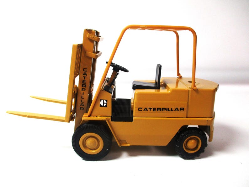 Może przedstawiać: Ż&oacute;łta zabawka w&oacute;zka widłowego Caterpillar z czarnymi oponami i czarnym siedzeniem. W&oacute;zek widłowy posiada mechanizm podnoszenia, a na boku widnieje napis "CATERPILLAR". Zabawka jest miniaturową repliką pojazdu budowlanego.