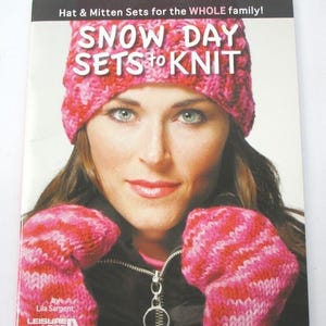 May include: A book cover featuring a woman wearing a pink and red knit hat and mittens. The title reads "SNOW DAY SETS to KNIT". The cover also includes the text "Hat & Mitten Sets for the WHOLE family!" and the author's name, Lila Sargent.