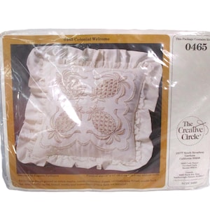 May include: A packaged pillow kit featuring a cream-colored pillow with embroidered pineapple designs. The pillow has a ruffled edge and is packaged in clear plastic. The package includes the text "0465 Colonial Welcome".