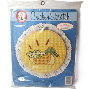 May include: A Chicken Scratch embroidery kit in a plastic package. The kit includes an 8-inch wooden hoop and a design featuring a duck in a basket with cattails. The design is on a yellow gingham background, surrounded by a white lace doily.