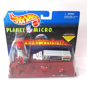 May include: Hot Wheels Planet Micro Armageddon toy set featuring a space shuttle, a semi-truck with a trailer, and two small figures on a red base. The packaging includes the text Hot Wheels, Planet Micro, and Armageddon with a space background.