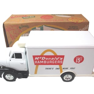 May include: A white and black McDonald's delivery truck toy with the McDonald's logo and text. The truck has a white cargo area with the text "McDonald's Hamburgers" and "There's one near you!". A cardboard box is behind the truck.