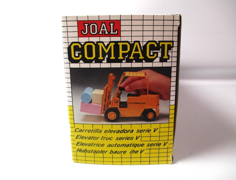 Może przedstawiać: Pudełko na zabawkę JOAL Compact. Pudełko przedstawia ż&oacute;łto-pomarańczowy w&oacute;zek widłowy podnoszący małe klocki. Na pudełku znajduje się napis "COMPACT" dużymi ż&oacute;łtymi literami. Pudełko zawiera r&oacute;wnież tekst w wielu językach.