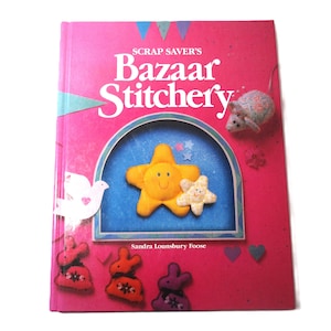 May include: A pink book titled "Scrap Saver's Bazaar Stitchery" with a blue and white illustration of a star and a smaller star. The cover also features a white dove, a mouse, and several felt bunny figures.