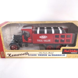 May include: Die-cast metal Kenworth stake truck with barrels. The red truck has black accents and the text "True Value" on the side. The truck is in a box with the text "1925" and "Kenworth STAKE TRUCK W/BARRELS".