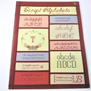 May include: A vintage book cover titled "Script Alphabets" by Henrietta Tour. The cover features various script alphabet examples in different colors and styles, including floral embellishments. The book is in a rectangular format.