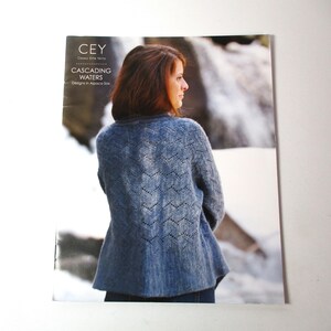 May include: A blue knitted cardigan with a lacy pattern, worn by a person standing in front of a snowy landscape. The text on the cover reads "CEY Classic Elite Yarns CASCADING WATERS Designs in Alpaca Silk."
