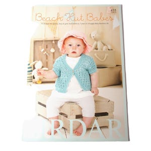 May include: A book cover featuring a baby wearing a light blue knitted cardigan and a pink hat. The book title is "Beach Hut Babies" and the subtitle is "14 designs for babies, boys & girls from birth up to 7 years in Snuggly Baby Bamboo DK."