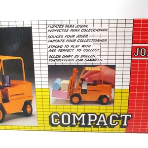 Może przedstawiać: Zabytkowe pudełko z zabawkami z pomarańczowym w&oacute;zkiem widłowym Caterpillar. Pudełko zawiera tekst w wielu językach, w tym &bdquo;Solidne do zabawy i idealne do kolekcjonowania&rdquo;. Widoczna jest nazwa marki &bdquo;JOAL&rdquo;.