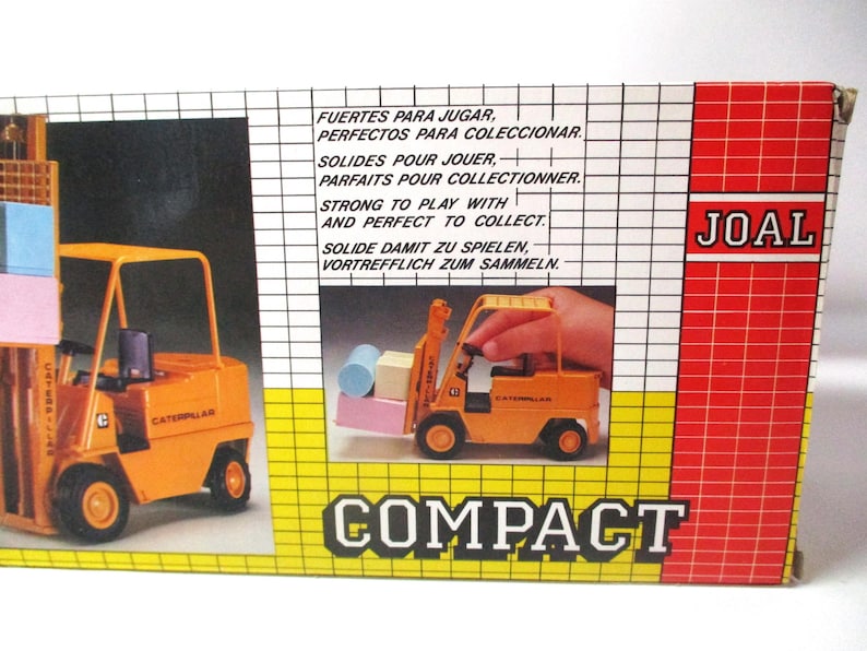 Może przedstawiać: Zabytkowe pudełko z zabawkami z pomarańczowym w&oacute;zkiem widłowym Caterpillar. Pudełko zawiera tekst w wielu językach, w tym &bdquo;Solidne do zabawy i idealne do kolekcjonowania&rdquo;. Widoczna jest nazwa marki &bdquo;JOAL&rdquo;.