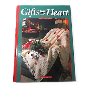 May include: A book titled "Gifts from the Heart" with a green cover and red spine. The cover features a photo of a cream-colored crocheted blanket with red floral accents draped over a chair. Decorative items are on the chair.