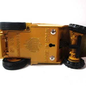 Może przedstawiać: Ż&oacute;łta zabawka, pojazd budowlany, do g&oacute;ry nogami, z czarnymi oponami. Sp&oacute;d zawiera napis "MADE IN SPAIN" i "N&ordm; 215 MINIATURAS". Zabawka ma szczeg&oacute;łowy design i jest wykonana z metalu.