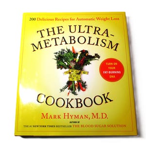 May include: A yellow cookbook titled "The Ultra-Metabolism Cookbook" by Mark Hyman, M.D. The cover features a bunch of vegetables tied with a measuring tape and a red circle with the text "Turn on your fat-burning DNA".