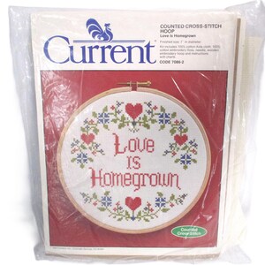 May include: A cross-stitch hoop kit in a plastic package. The design features the words "Love Is Homegrown" in red script, surrounded by hearts and floral accents. The finished size is 7 inches in diameter.
