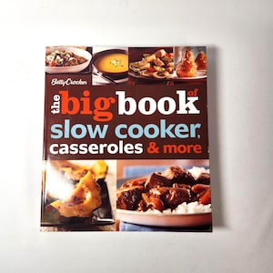 May include: A cookbook titled "The Big Book of Slow Cooker, Casseroles & More" by Betty Crocker. The cover features images of various cooked dishes, including soups, casseroles, and meat dishes. The book is set against a white background.