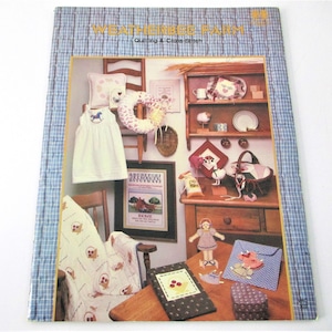 May include: A book cover titled "Weatherbee Farm" with a blue plaid border. The cover features a collage of handmade crafts, including a white dress with a horse applique, a wreath, and a framed sampler. A wooden shelf displays decorative items.