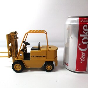 Może przedstawiać: Mała, ż&oacute;łta zabawka w&oacute;zka widłowego Caterpillar z czarnymi kołami i czarnym siedzeniem. W&oacute;zek widłowy stoi obok puszki Diet Coke.