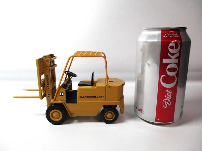 Może przedstawiać: Mała, ż&oacute;łta zabawka w&oacute;zka widłowego Caterpillar z czarnymi kołami i czarnym siedzeniem. W&oacute;zek widłowy stoi obok puszki Diet Coke.