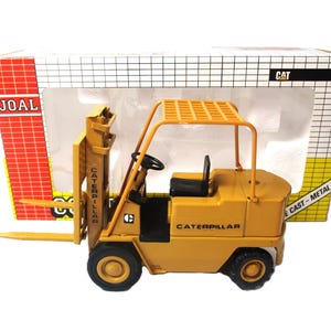 Może przedstawiać: Ż&oacute;łta zabawka w&oacute;zka widłowego Caterpillar z odlewanego metalu. W&oacute;zek widłowy ma czarne opony, czarne siedzenie i kierownicę. Pudełko ma logo JOAL i CAT.