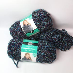 May include: Three skeins of Country Loom yarn in shades of teal, purple, and black. The yarn is thick and textured, with a label featuring a woman wearing a knitted scarf. The label also includes the text "Telas de Campestre".