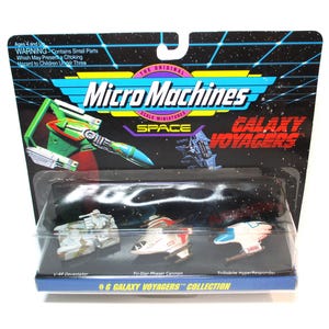 May include: A Micro Machines Space Galaxy Voyagers collection featuring three miniature spaceships. The spaceships are a gray and white U-48 Destroyer, a white and blue Tr-Star Phaser Cannon, and a white and blue Trilobite HyperResponder. The packaging is black with a red and yellow Micro Machines logo and a red and white Galaxy Voyagers logo.